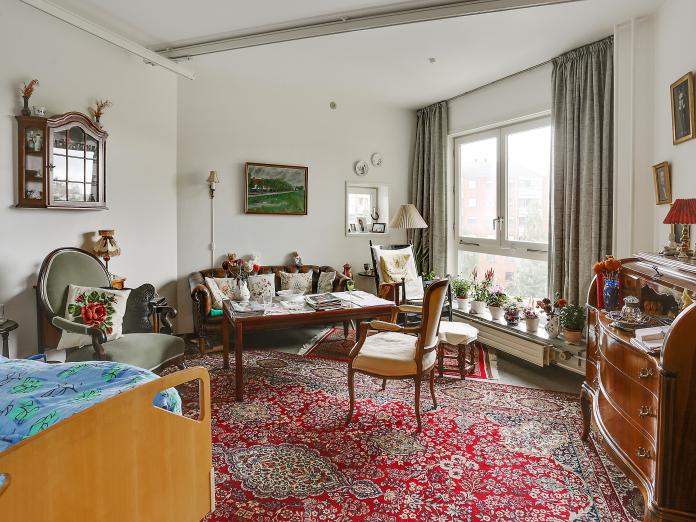 Lyst værelse med plejeseng, loftslift og personlige møbler som sofa, lænestole, sofabord, chatol og gulvtæpper.