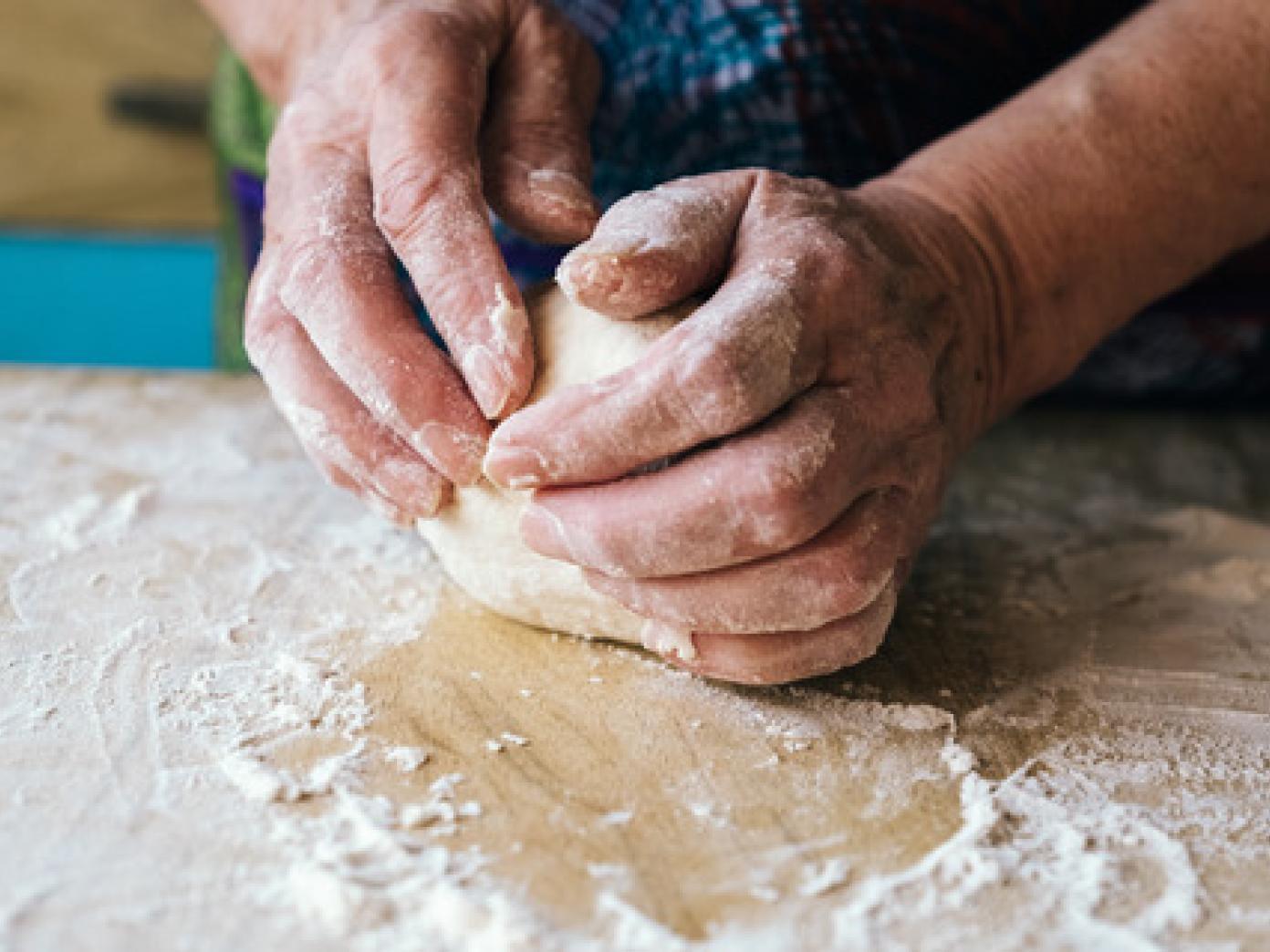 Nærbillede af hænder dækker af mel, der ælter en dej på et bord drysset med mel.