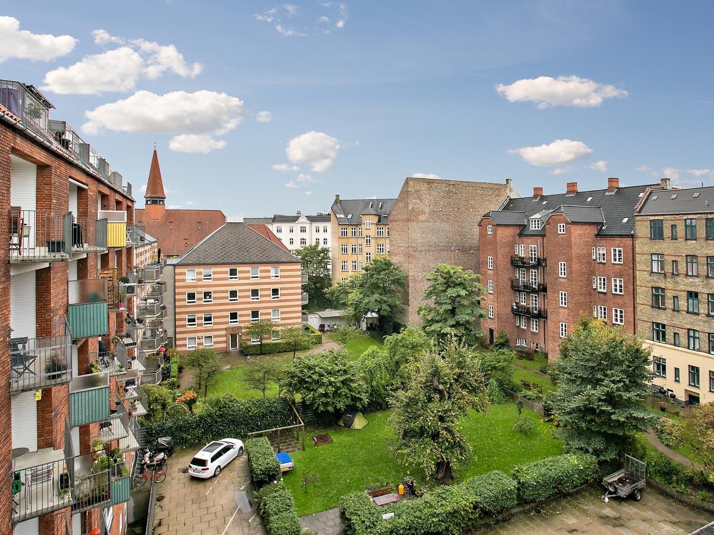 Udsyn over grøn gårdhave med hække, buske, træer og bede. Gårdhaven er omkranset af murstens etagebyggerier af ældre dato og nyere dato.