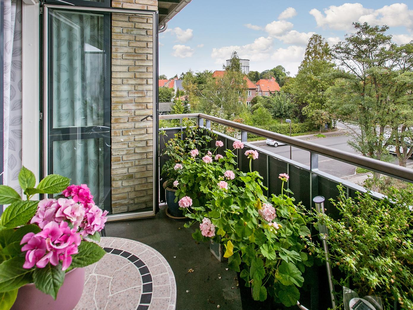 Privat altan med blomster i krukker og kig ud mod vej med høje træer.