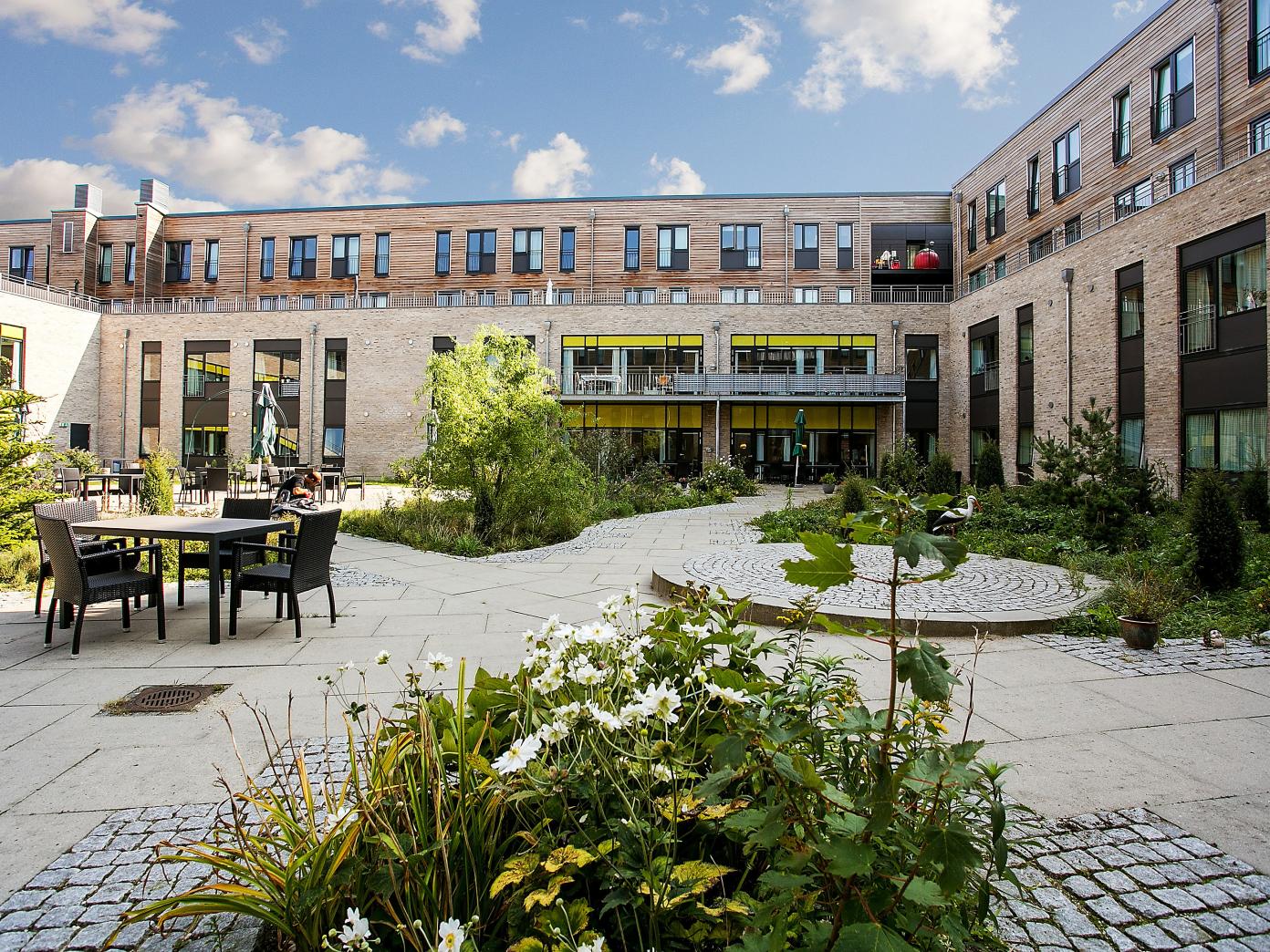 Indre gårdhave omkranset af en moderne bygning i lyse sten og træ. Gårdhaven er indrettet med bede, buske og træer samt gangstier og terrasser med havemøbler.