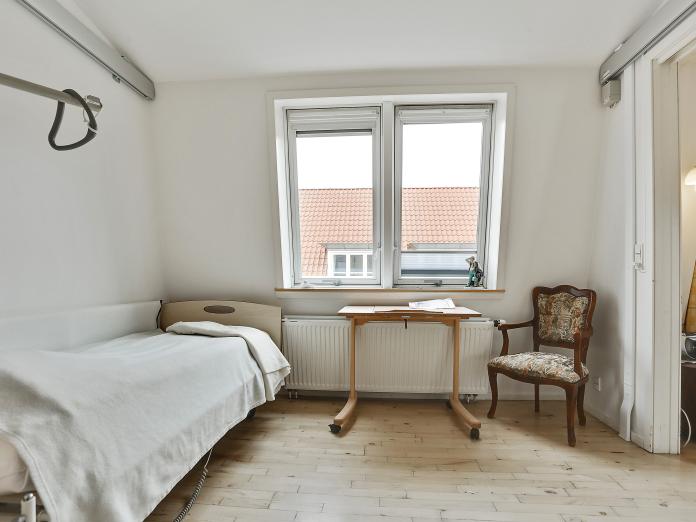 Soveværelse med plejeseng, skråvinduer, lille bord og stol. I loftet er der skinner til loftlift.