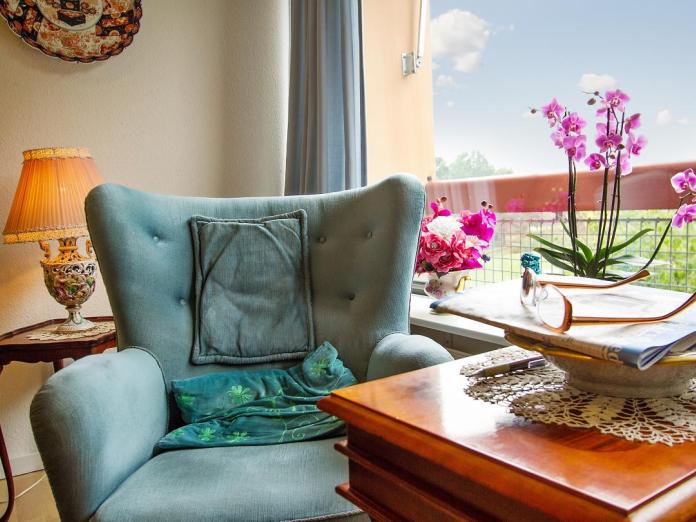 Grøn lænestol og små konsolborde i mørkt træ foran stort vindue med blomster i vindueskarmen. 