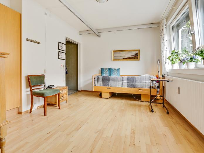 Soveværelse med skab, plejeseng, stol og skinner til loftlift. I vindueskarmen er potteplanter.