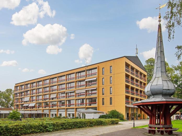 Gult etagebyggeri med sorte vinduer, franske altaner og markiser samt en park med pavilloner, træer og buske. I forgrunden ses et spir i træ og kobberplader med en bænk nedenunder. 