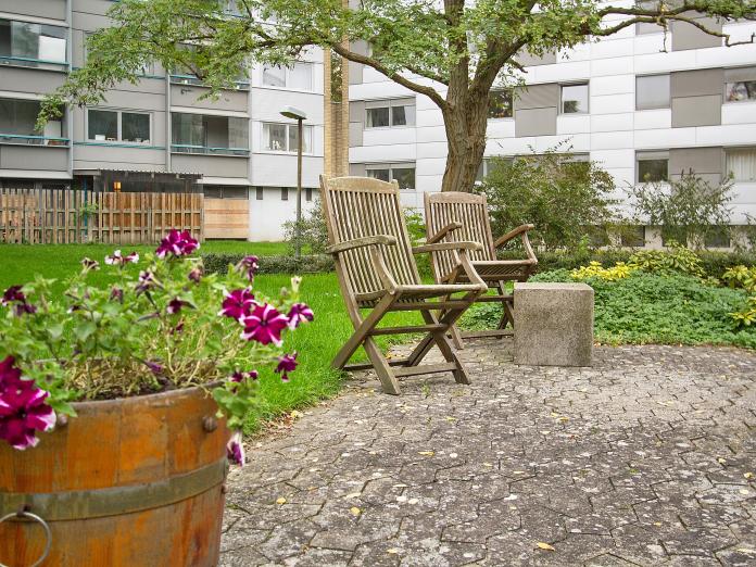 Havemiljø med terrasse, blomster, træer, et stakit i baggrunden og to havestole i træ.