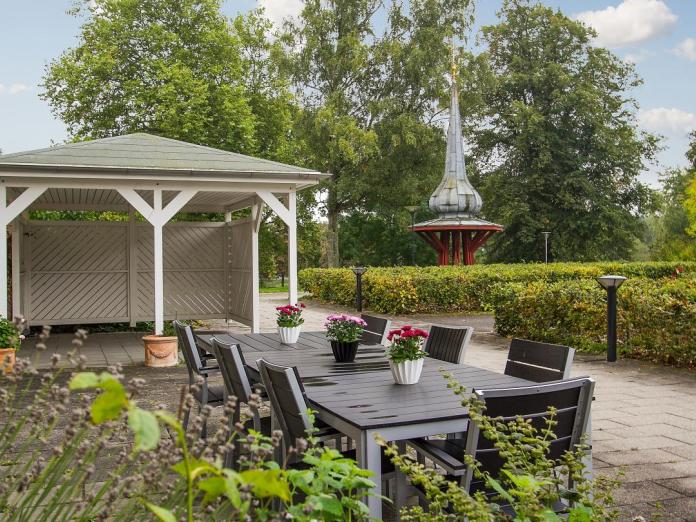 Grøn gård med haveborde og stole, grønne, trimmede hække, høje træer, pavillon, krukker med blomster og et spir påført i træ og kobber.