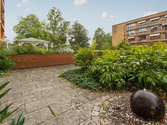 Gårdhave med fliser, grønne buske, højbede med urter, høje træer samt lille springvand.