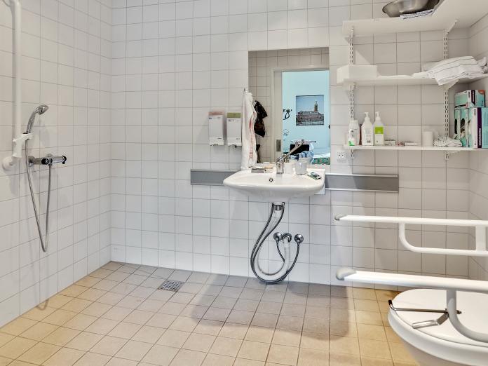 Hvidt flisebadeværelse med vask, spejl, toilet og bruser samt hylder til personlige plejeprodukter.