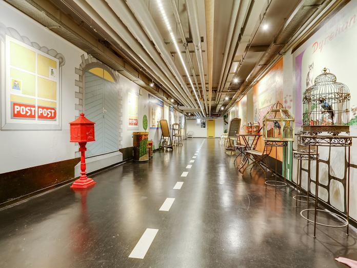 Lang gang indrettet som gammel handelsgade med caféstole og bord, rød postkasse, striber på gulvet og butiksfacader malet på væggen.