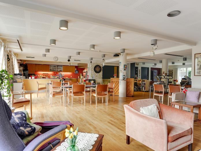 Stort fællesrum med lænestole, en række borde med stole, blomster, planter, et klaver og et køkken.