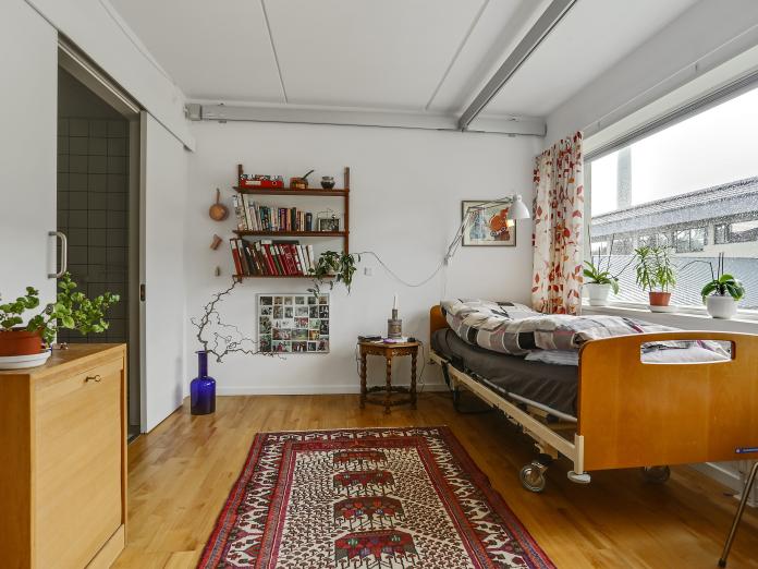 Soveværelse med seng, reoler og billeder på væggene, tæppe på gulvet og skydedør til badeværelse.