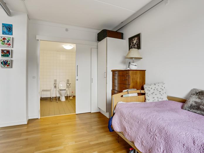 Lyst, soveværelse med plejeseng, indbygget skab og kommode i mørkt træ. Soveværelset har også en loftslift samt adgang gennem skydedør til badeværelse.