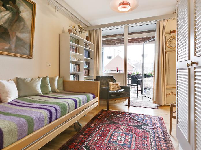 Soveværelse med stort vinduesparti samt dør ud til privat altan. Soveværelset er indrettet med plejeseng, bogreol, stole og skab. Derudover hænger der et maleri på væggen og på gulvet ligger et gulvtæppe.