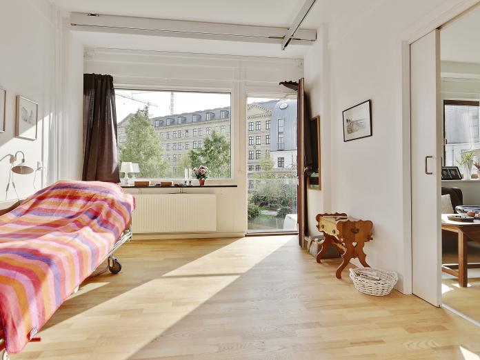 Lyst soveværelse med plejeseng, loftslift og stort vindue samt fransk altan. Værelset er indrettet med personlige møbler og malerier på væggene.