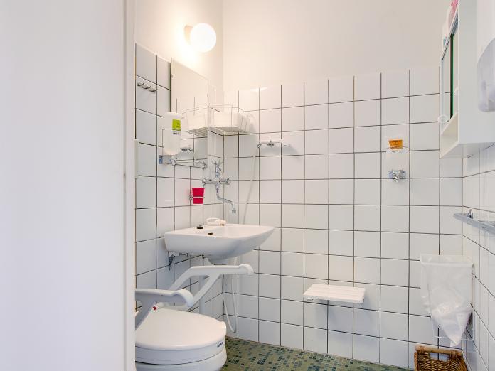 Hvidt flisebadeværelse med vask, spejl, toilet med armstøtter samt bruser og brusebænk.