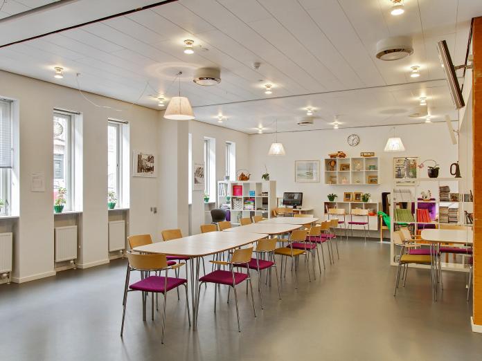 Lyst, aflangt fællesrum indrettet med langbord med stole omkring samt reoler med nips og grønne planter.