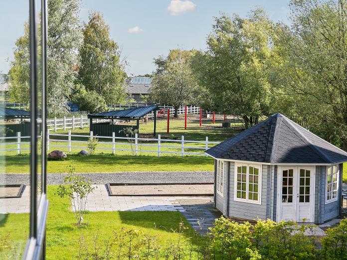 Udsigt fra bygning ned mod have med græs, buske og træer samt orangeri. I det fjerne er en fold til heste.