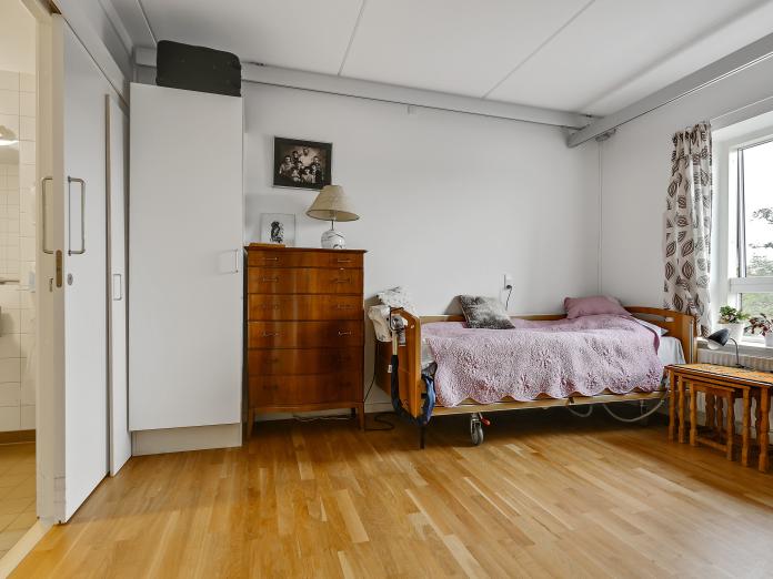 Lyst soveværelse med plejeseng, indbygget skab, loftslift og stort vindue. Fra soveværelset er der adgang til et badeværelse.