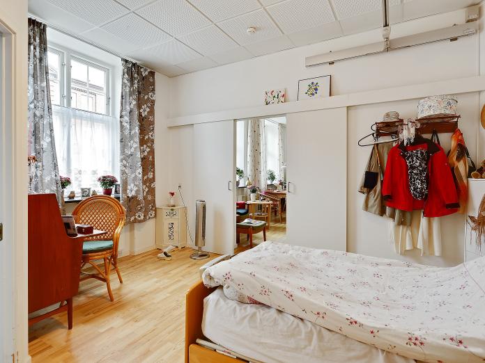 Lyst soveværelse med plejeseng og loftslift samt skydedøre ind til stue. Værelset er møbleret med personlige møbler og billeder på væggene.