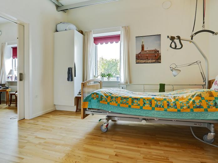 Lyst soveværelse med trægulve indrettet med plejeseng, loftslift og indbygget skab. Der er en skydedør mellem soveværelse og stue.