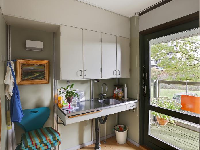 Lyst tekøkken med overskabe og bordplade med vask. Fra tekøkkenet er der en dør ud til altan. 