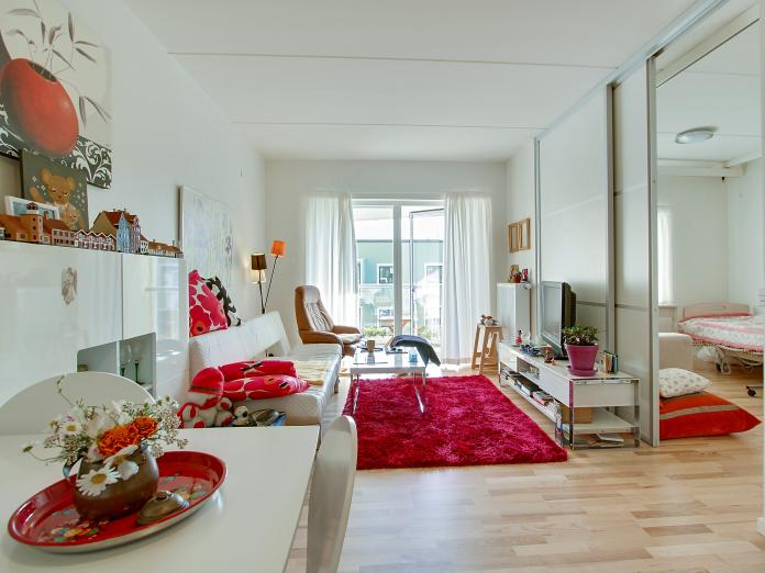 Lyst værelse med stort vindue og dør ud til altan. Værelset er indrettet med personlige møbler som sofa, lænestol, reolsystem, fjernsyn og et spisebord med stole omkring.