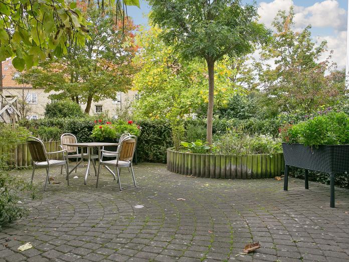 Lille grøn, krog i gårdhaven med høje træer, højbede med blomster og urter samt fire stole omkring et cafébord. 