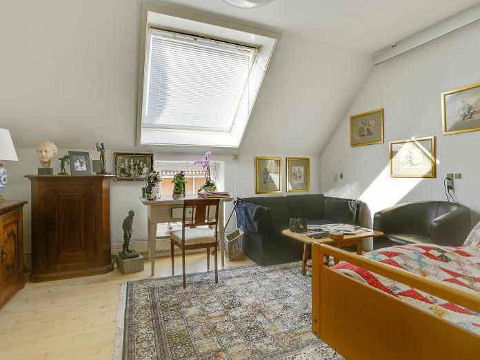 Lyst værelse med ovenlysvindue, plejeseng og loftslift. Værelset er møbleret med personlige møbler som sofa, lænestol og sofaborde samt kommoder og et skrivebord med stol.