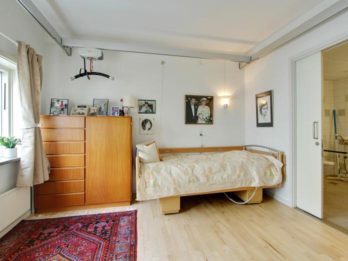 Lyst soveværelse med plejeseng, loftslift og personlige møbler som chatol og billeder på væggene. Fra soveværelset er der adgang til badeværelse.