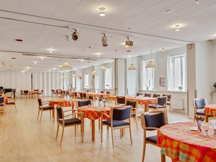 Stort, lyst fællesrum med borde med dug på og stole omkring.