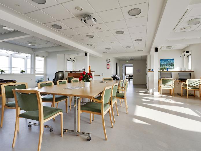 Stort, lyst fællesområde med langbord og stole, to computerstationer, lænestole, sofaer og et klaver.