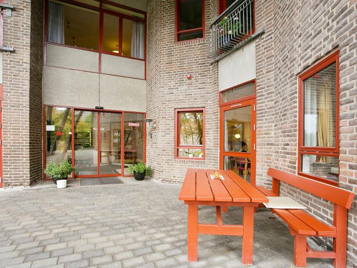 Udeareal foran indgangen til bygningen. Her er et bord og en bænk samt grønne planter i krukker foran døren. 