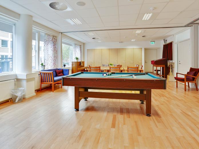 Lyst fællesrum med billardbord, klaver, sofa og lænestole samt langbord med stole omkring.