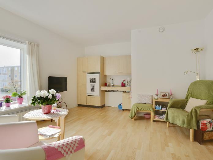 Lys stue med tekøkken og indrettet med personlige møbler som læsestole, sideborde og et fjernsyn.