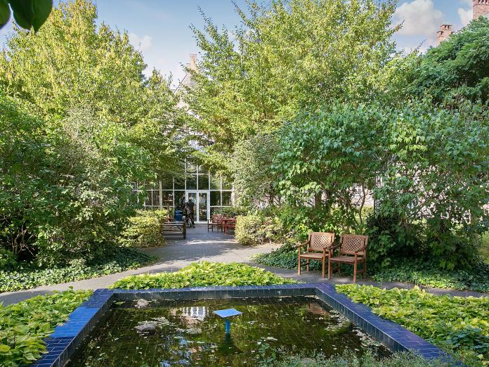 Gårdhave med gangstier, bede, buske og høje træer, i forgrunden er et havebassin i blå fliser. Haven er indrettet med havemøbler i mørkt træ.