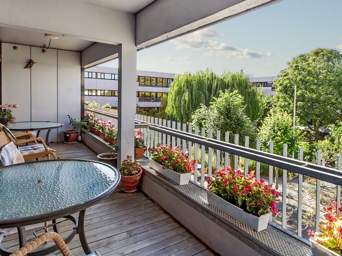 Fælles altan indrettet med caféborde og kurvestole samt altankasser med blomster og blomster i krukker. Fra altanen er der kig udover en gårdhave med buske og store træer.