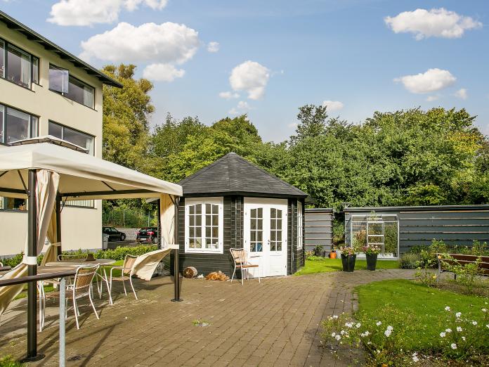 En terrasse, græsplæne, forskellige bed, et sort tehus med hvide vinduer og døre, en pavillon og forskellige havemøbler. I baggrunden et stort trætæppe fra Lersøparken.