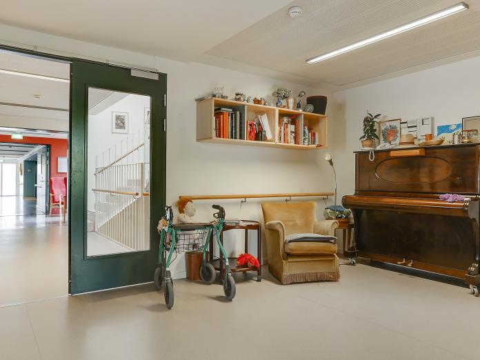 Fællesrum med en parkeret rollator, bogreol, lænestol og et klaver med mange nips ovenpå.