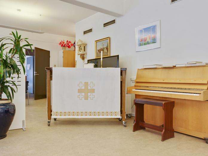 Fællesrum med klaver og højbord med alterdug. På væggene hænger billeder og på gulvet står en stor, grøn plante i en krukke.