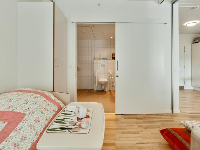 Soveværelse med plejeseng, loftslift og indbygget skab. Fra soveværelset er der adgang til stue med tekøkken og til et stort flisebadeværelse.