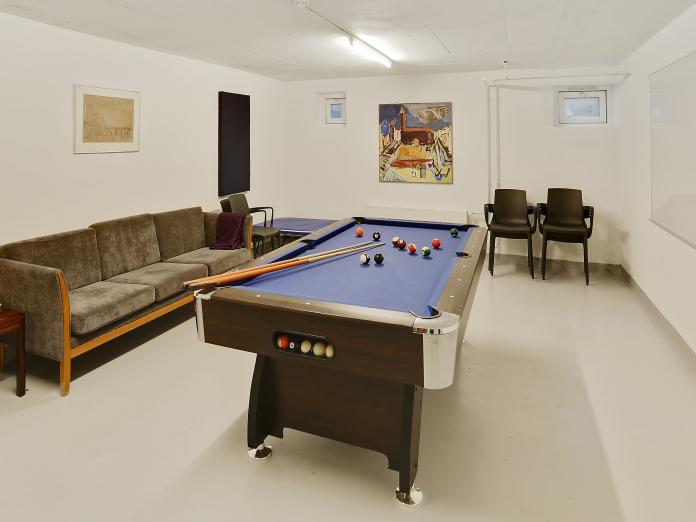 Lyst kælderlokale med billardbord, sofa, stole og kunst på væggene.