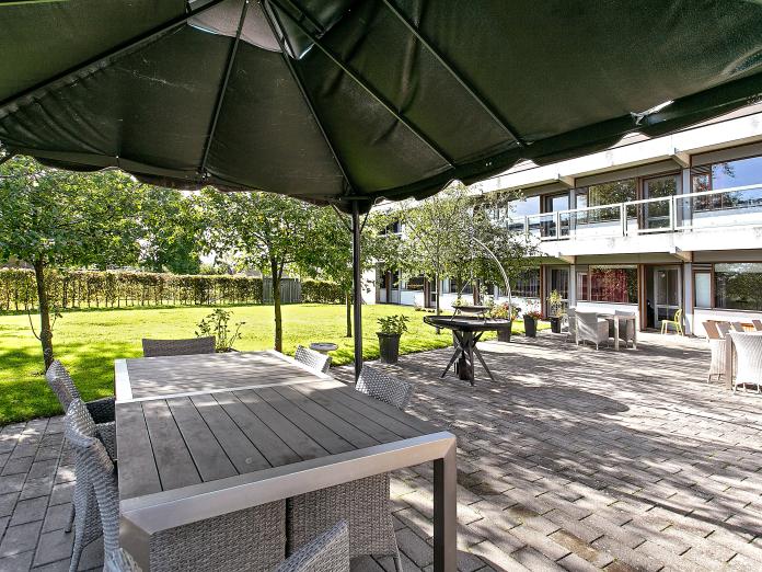 Fliseterrasse med havemøbler, krukker med blomster og en parasol samt areal med grønt græs og træer. I baggrunden en grå bygning med svalegang.