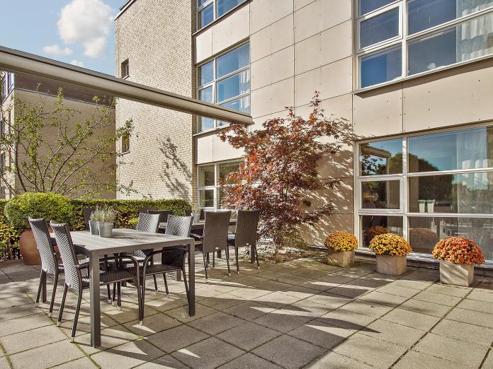 Fliseterrasse med havemøbler, træer og blomster i krukker samt en stor markise.