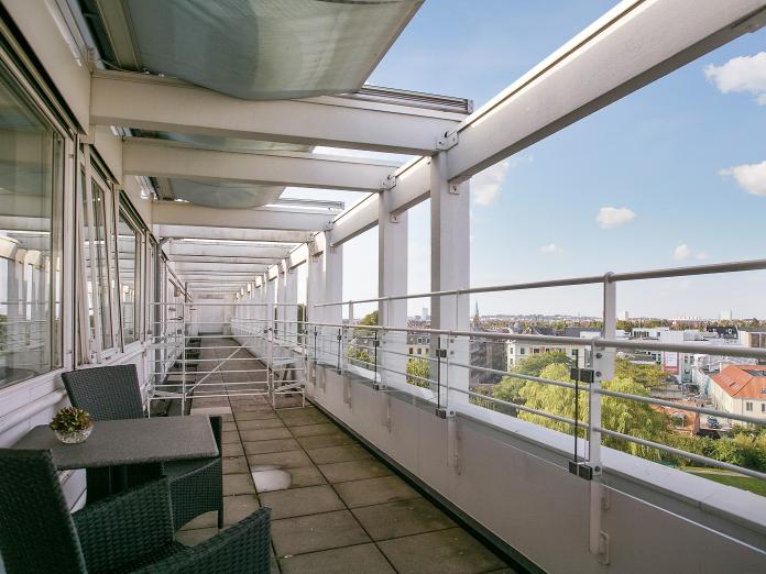 Fælles fliseterrasse med markiser og udsigt over Nørrebro. Terrassen er indrettet med kurvemøbler og caféborde med små potteplanter.   