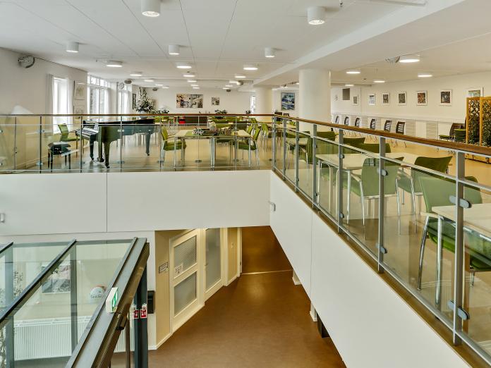 Åben førstesal med flygel, borde og stole samt grønne planter, hvorfra der er kig ned til indgangsparti.