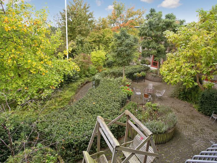 Grøn gårdhave med trimmede hække, højbede med blomster og krydderurter, høje træer samt borde, stole og en hængesofa. 