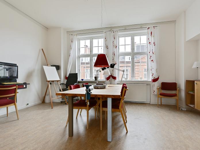 Lyst lokale indrettet til kreative aktiviteter med bord og stole samt et staffeli i hjørnet og et computerbord.