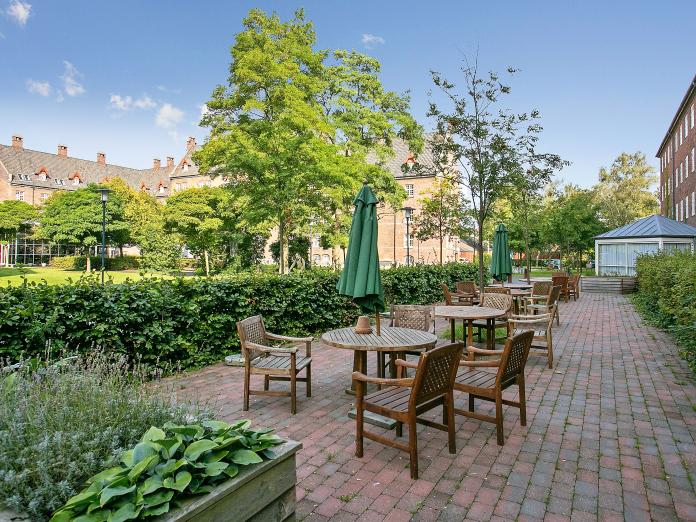 Fliseterrasse omkranset af bøgehække og indrettet med højbede med grønt og urter samt havemøbler i mørkt træ og parasoller.