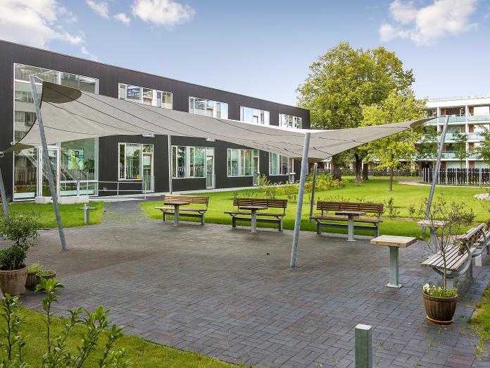 Terrasse med bænke, krukker og en græsplæne i baggrunden. På terrassen er et stort, hvidt solsejl.